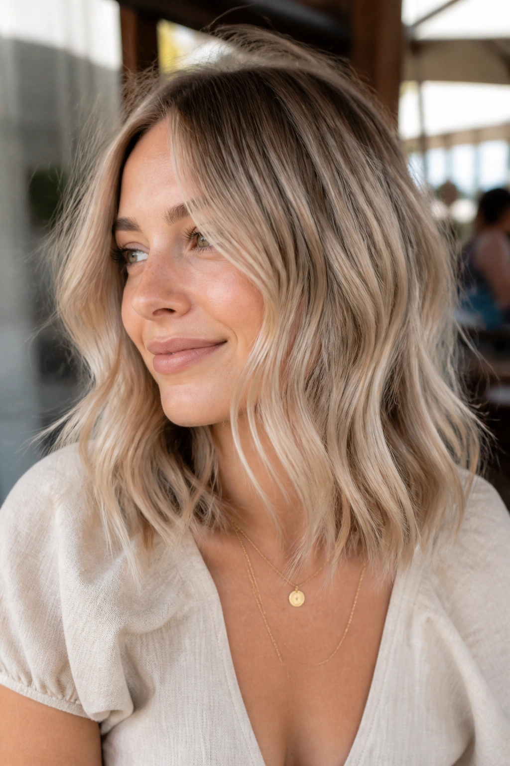 Medium close-up of a real woman with beige blonde waves in a lob cut in sunny outdoor cafe light.