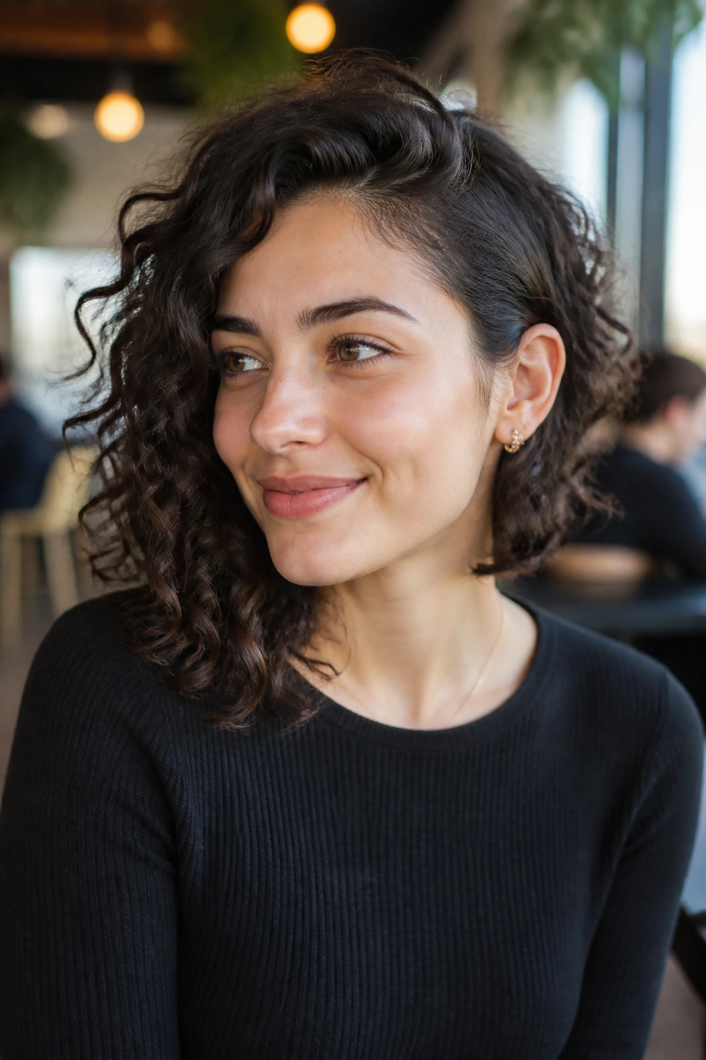 Medium-close portrait of a real person with collarbone-grazing curly lob and asymmetric length in cafe daylight