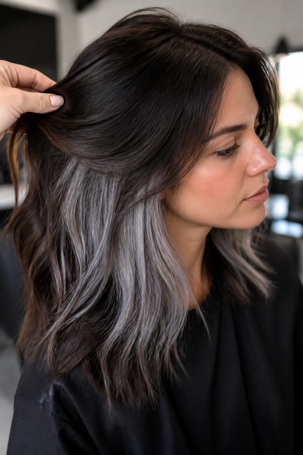 Close-up of a real woman showing hidden silver layers under dark brown hair at the crown in a modern salon.