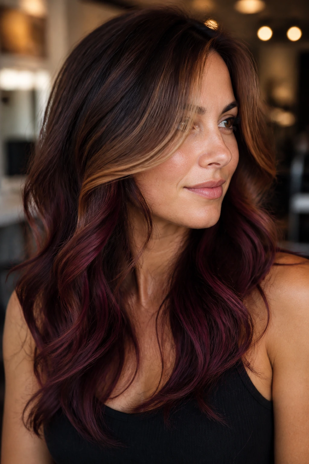Close-up portrait of a real woman with plum and caramel balayage on layered hair in a warm salon.